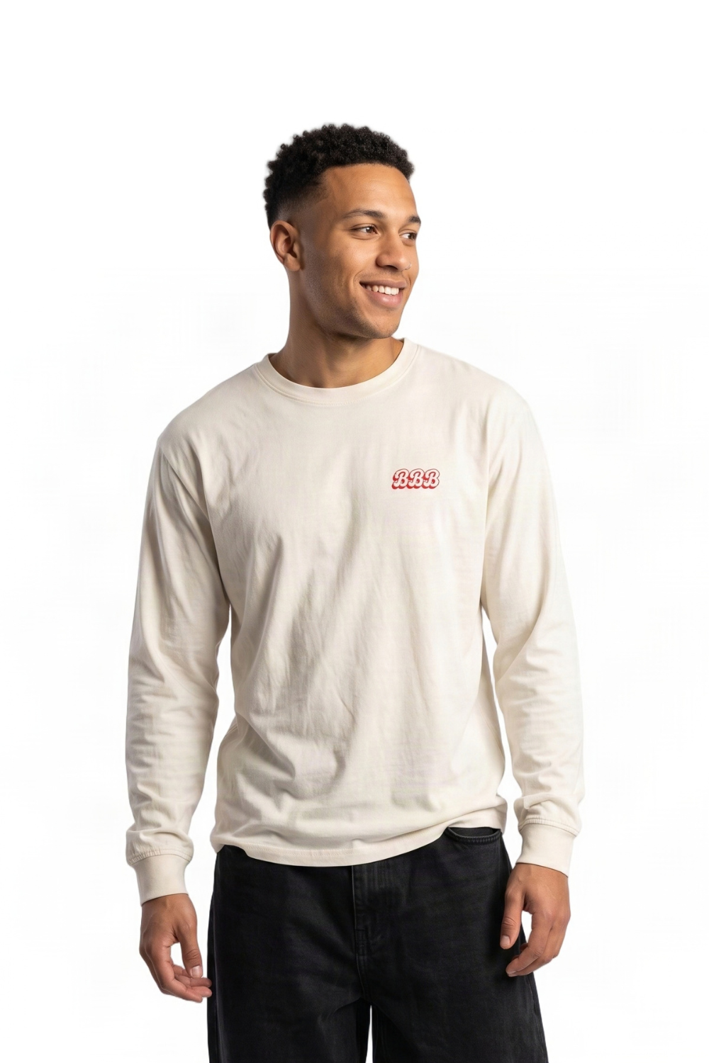 A man wearing a cream-colored long-sleeve crewneck sweatshirt with a small red embroidered logo on the left chest, paired with black jeans, against a plain white background.